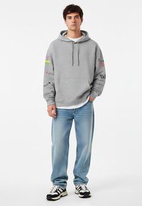 Sudadera gris con cordón, diseños gráficos en las mangas, combinada con jeans azul claro y zapatillas en blanco y negro. Tejido suave, corte casual.