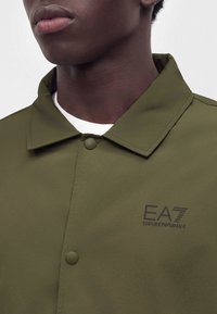 Крупен план на човек, облечен с маслинено зелено яке Emporio Armani EA7 с копчета на яката и бяла риза под него.