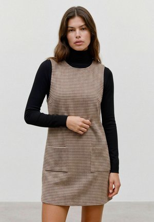 Femme aux cheveux bruns ondulés porte un haut noir à manches longues sous une robe marron et beige sans manches à motif pied-de-poule avec poches devant.