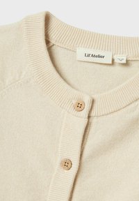 Gilet en tricot beige avec un col rond, deux boutons en bois et des poignets côtelés. Le tissu a une finition douce et texturée.