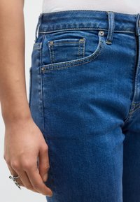 Σκούρο μπλε τζιν με εφαρμοστή σχεδίαση, διακοσμημένο με χρυσές ραφές, μικρή μπροστινή τσέπη και τυπικό κούμπωμα με κουμπί και φερμουάρ.