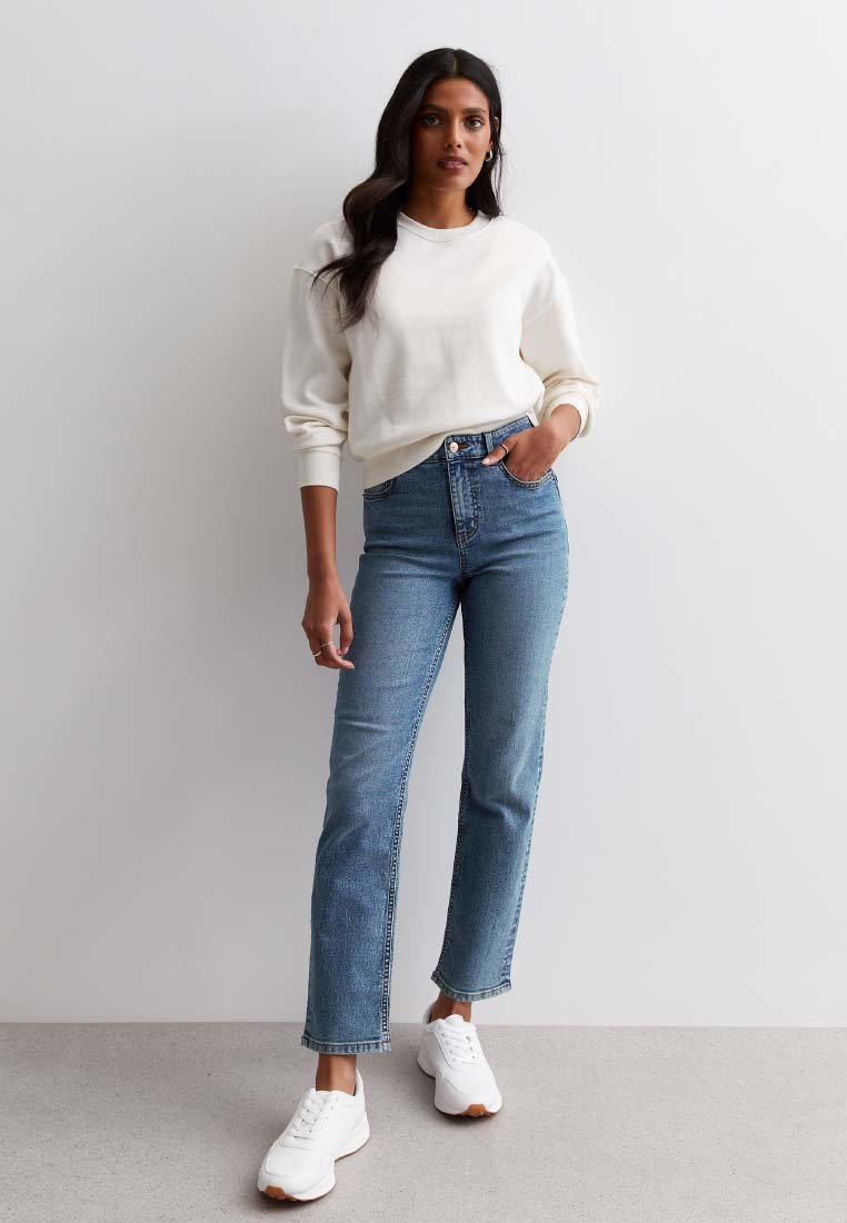 Vit sweatshirt med långa ärmar, blå högmidjade jeans och vita sneakers med en strukturerad sula, stående mot en ljus bakgrund.