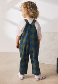 Une salopette à motifs losanges bleus et verts avec des bretelles, associée à une chemise blanche et des baskets blanches portées par un enfant.