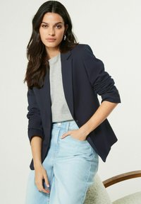 Námořnický blazer s ohrnutými rukávy přes šedé tričko, v kombinaci s světle modrými džíny; hladká látka, upravený design a detail přední kapsy.