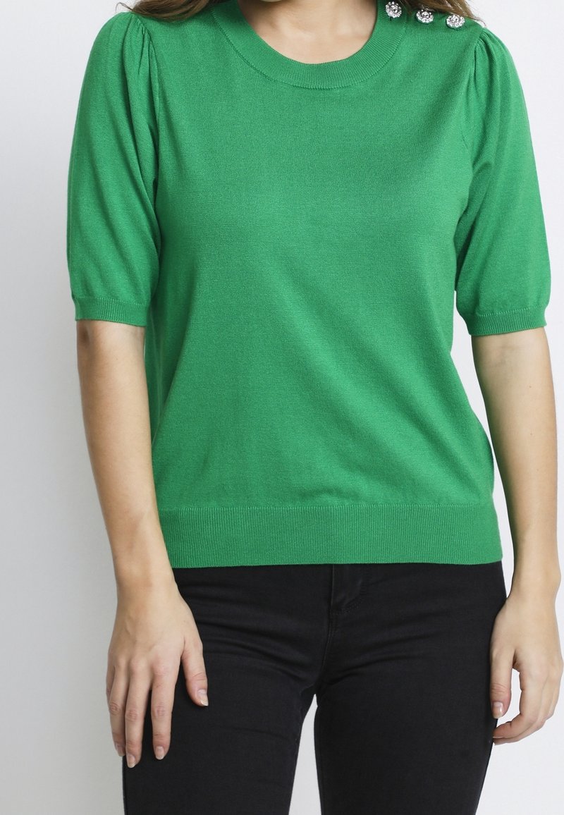Femme portant un pull vert à manches courtes avec des boutons décoratifs sur l'épaule et un pantalon noir, debout devant un fond uni.