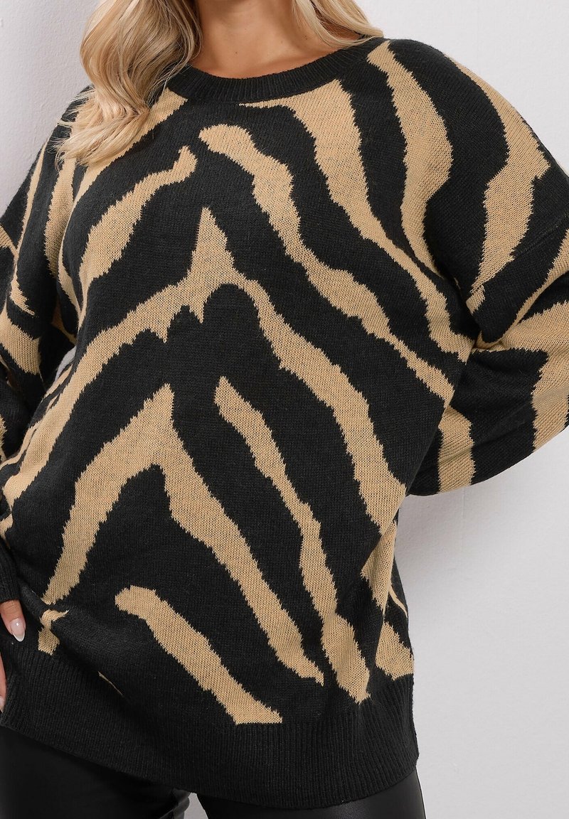 Femme portant un pull rayé zèbre noir et beige à manches longues avec poignets côtelés, posant devant un fond clair uni.