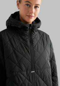 Zwarte gewatteerde pullover met een hoge kraag en capuchon. Voorzien van een ritssluiting aan de voorkant en een horizontale naad met een klein logoaccent.
