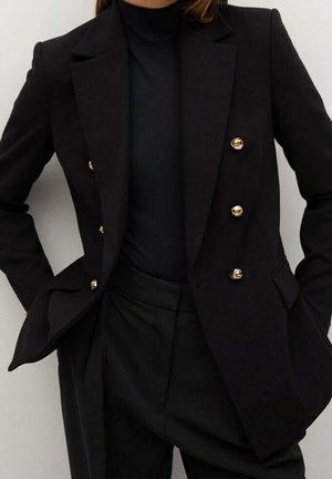 Czarna dwurzędowa marynarka z złotymi guzikami, noszona na czarnych, wysokich spodniach z wysokim stanem i czarnym golfie, z rękami w kieszeniach.