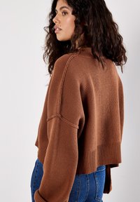 Brown, knitted cropped sweater with ribbed hem and sleeve cuffs. Features a relaxed fit and textured pattern. Paired with blue jeans.