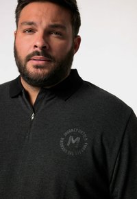 Schwarz mit einem halben Reißverschluss, aus strukturiertem Stoff und mit einem kreisförmigen Logo-Detail auf der Brust.