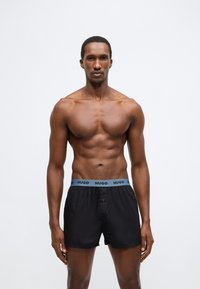 Μαύρα ανδρικά εσώρουχα μπόξερ με ανοιχτό μπλε ελαστική μέση που φέρει το λογότυπο "HUGO". Το υλικό φαίνεται λείο και ελαφρύ.