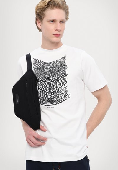 Balta T-kreklu ar melnu līniju grafisko dizainu, apvienots ar melnu plecu somu ar rāvējslēdzēja kabatu un regulējamu siksnu.