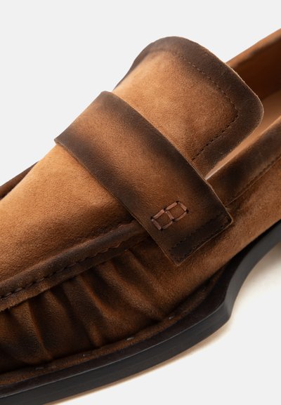 Καφέ μοκασίνι από σουέτ με ραμμένο λουράκι και πτυχώσεις πάνω από τη μαύρη σόλα, σε κοντινή γωνία σε λευκό φόντο.