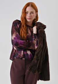 Donna con lunghi capelli rossi che indossa una blusa viola con un motivo e pantaloni bordeaux, mentre porta un cappotto di pelliccia marrone scuro sulle spalle.