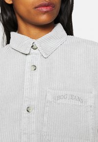 Kobieta nosząca jasno szary prążkowany koszulowy sweter z guzikami i kieszenią na piersi wyszywaną napisem "BDG JEANS."