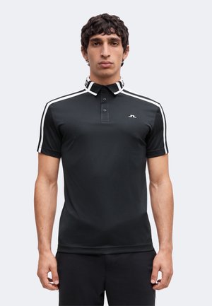 Man draagt een zwarte polo met korte mouwen met witte strepen op de mouwen en kraag, staand met het gezicht naar voren tegen een effen achtergrond.