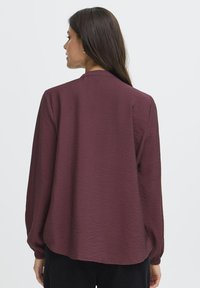 Femme aux longs cheveux noirs, portant une blouse ample à manches longues couleur bordeaux, se tenant de dos contre un fond clair.