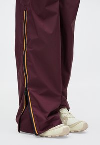 Spodnji deli noge nosijo bordo hlače z delno odprtimi stranskimi zadrgami, ki razkrivajo bež in bele športne čevlje na beli podlagi.