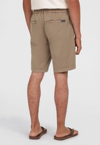 Beige Shorts mit elastischem Bund, zwei Gesäßtaschen und einem Logo-Tag. Glattes Material mit lässigem Schnitt, kombiniert mit braunen Flip-Flops.