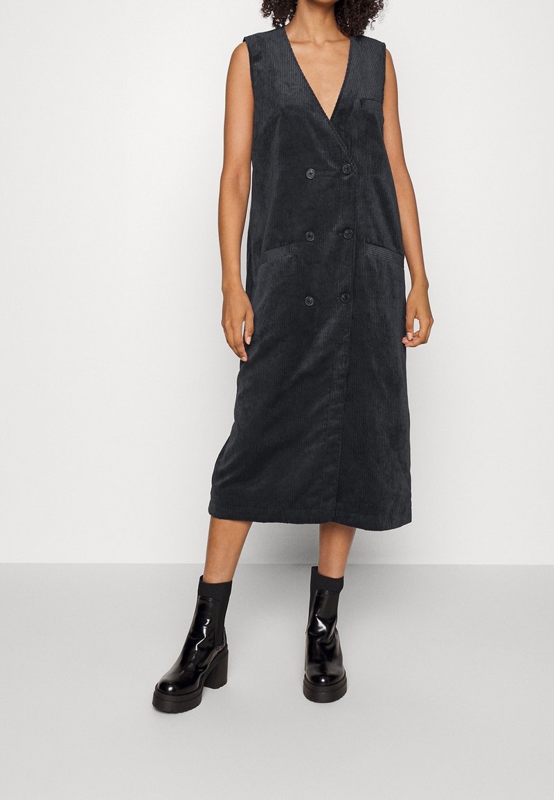 Nainen, joka käyttää mustaa hihattomasta corduroy-mekkoa, jossa on kaksiriviset napit, yhdistettynä mustiin korollisiin nilkkureihin.