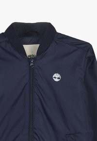 Giubbotto bombardiere blu navy con colletto a coste, chiusura con zip e un piccolo logo bianco di un albero sul lato sinistro del petto. Tessuto liscio, design leggero.
