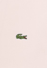 Logo de cocodrilo bordado en verde con la boca roja abierta sobre una tela texturizada de color rosa claro.