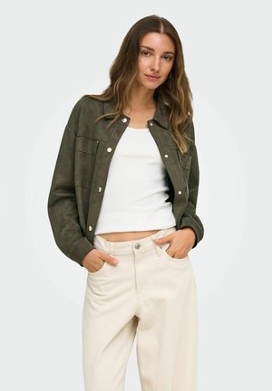 Femme aux longs cheveux ondulés porte une veste vert foncé sur un top court blanc et un pantalon taille haute beige clair, debout les mains dans les poches.