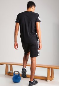 Muž v černém sportovním oblečení stoupá na dřevěnou lavici s modrou kettlebell na podlaze poblíž, proti jednoduché zdi.