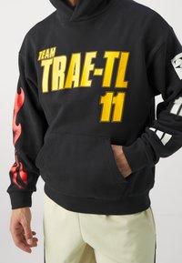 Hoodie noir avec grand texte jaune "TEAM TRAE-TL" et "11." Motifs de flammes rouges sur les manches, la poche avant et les poignets côtelés.