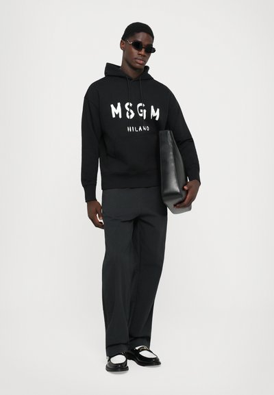 Uomo che indossa una felpa nera MSGM Milano, pantaloni neri, mocassini bianco e neri, occhiali da sole scuri, mentre tiene una grande borsa di pelle nera.