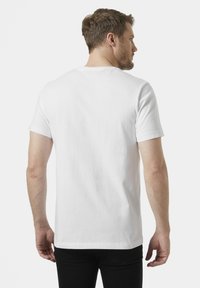 Camiseta de algodón blanca con cuello redondo y mangas cortas, vista desde la parte posterior. Textura suave, corte estándar. Jeans negros visibles por debajo.