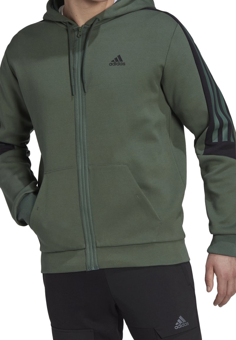 Homme portant un hoodie zippé vert Adidas avec des bandes noires sur les manches et un pantalon noir Adidas avec logo sur la cuisse.