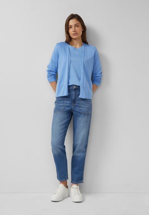 Frau steht vor einer grauen Wand, trägt eine hellblaue Strickjacke, ein passendes T-Shirt, blaue Jeans und weiße Sneaker, die Hände hinter dem Rücken verschränkt.