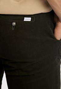 Donkergroene corduroy broek met fijne ribbeltextuur, functionele zakken en een knoopsluiting met een blauwe knoop en een label op de tailleband.