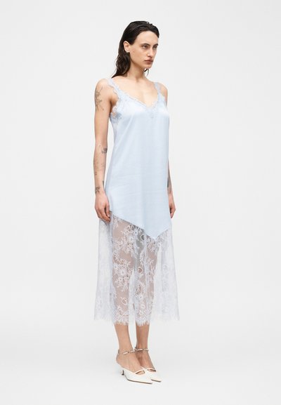 Γυναίκα με τατουάζ που φοράει ανοιχτό μπλε αμάνικο φόρεμα με δαντέλα στο τελείωμα και λευκές μυτερές γόβες με λουράκια στον αστράγαλο, σε λευκό φόντο.