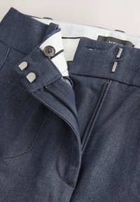 Pantalon en denim bleu foncé avec braguette partiellement ouverte, fermeture par bouton et attaches métalliques à crochet et barre sur la ceinture.