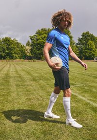 Maillot de sport bleu avec des accents bleu clair, short noir, chaussettes blanches hauteur genou et chaussures de sport blanches ; tenant un ballon de football sur un terrain enherbé.