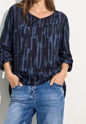 Femme portant un haut à manches longues bleu foncé à motifs avec les manches retroussées et un jean bleu clair, les mains dans les poches.