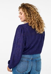 Tumši zila blūze ar mežģīnēm, pūkainām piedurknēm un brīvu piegriezumu. Valkāta kopā ar gaiši ziliem džinsiem, kas izceļ teksturētu auduma rakstu.