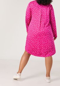 Robe fuchsia avec un motif de cœurs, manches longues et ourlet arrondi. Portée avec des baskets blanches, mettant en valeur une silhouette décontractée.