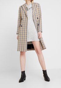 Cappotto trench doppiopetto in giallo, blu e bianco a quadri. Presenta un colletto e bottoni neri, abbinato a un vestito grigio.