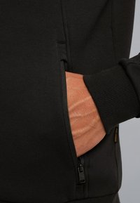 Sudadera negra con un bolsillo lateral, puños acanalados y textura suave. El detalle con cremallera en el bolsillo complementa el diseño minimalista.
