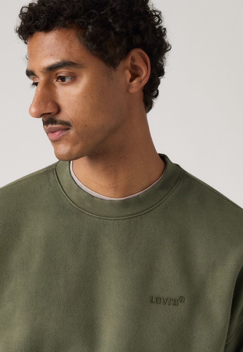 Giovane uomo con capelli ricci e baffi, indossa una felpa Levi's verde oliva, guardando verso sinistra su uno sfondo uniforme.