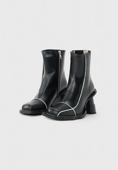 Μαύρα δερμάτινα μποτάκια αστραγάλου με λευκή ραφή, πλαϊνές φερμουάρ, φαρδιά τακούνια και στρογγυλεμένες μύτες πάνω σε απλό ανοιχτό φόντο.