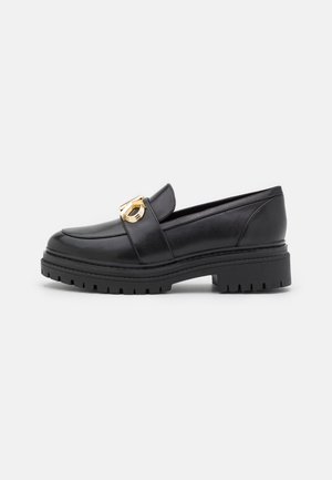 Mocassin en cuir noir avec semelle épaisse crantée et détail de boucle en métal doré sur le dessus, photographié sur un fond clair uni.