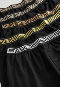 Schwarze Boxershorts mit gemusterten Gummibündchen in Gold, Silber und Schwarz. Jedes Bündchen zeigt ein griechisches Schlüsselmotiv. Weicher Stoff. Mit Knopfdetaillierung.