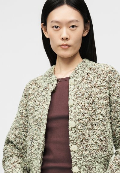 Jeune femme aux cheveux noirs et raides porte un cardigan en maille texturée crème et marron par-dessus un haut marron, regardant droit devant avec une expression neutre.