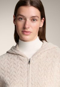 Krémově zbarvená pletená mikina s kapucí a copánkovým vzorem, s předním zipem a měkkým, texturovaným povrchem. Nošena přes bílý rolák.