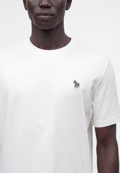 Homme portant un t-shirt blanc uni à manches courtes avec un petit logo zèbre coloré sur la poitrine gauche sur un fond uni.