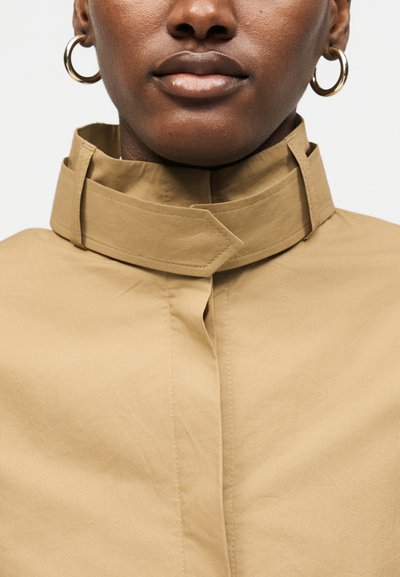 Gros plan d'une personne portant une veste beige à col montant avec des boucles d'oreilles en forme d'anneaux dorés sur un fond clair uni.
